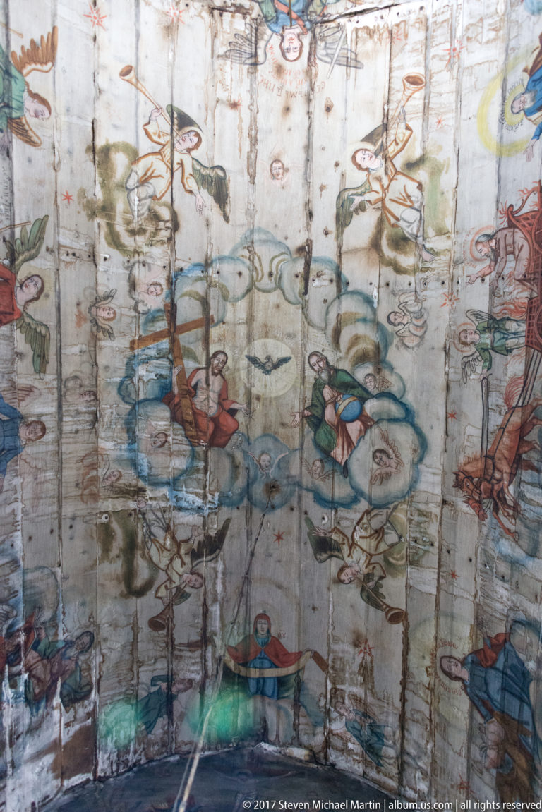 Faded religious scenes with figures, animals, and clouds painted across the curved plank ceiling of a wooden church.