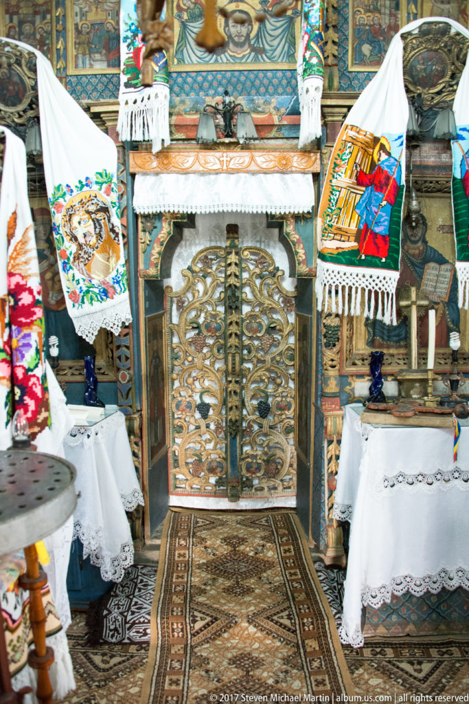 Carved iconostasis with painted icons, embroidered towels, lace-covered tables, and patterned rugs in a wooden church.