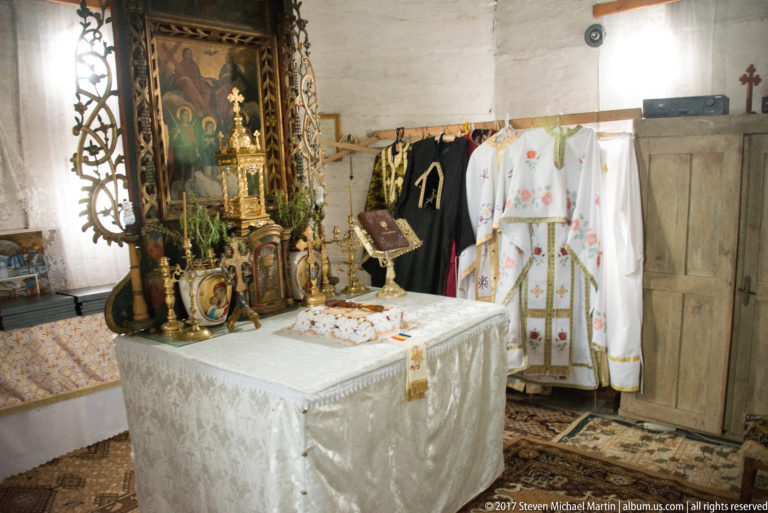 White-covered altar table beside carved church furnishings and hanging embroidered liturgical vestments.