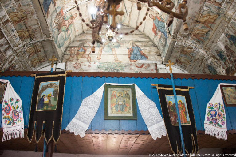 Painted wooden vault above a blue partition with framed icons, embroidered towels, chandelier, and processional objects.