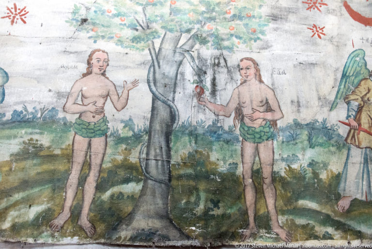 Painted Adam and Eve standing beside a tree, with Eve holding a red fruit, on a wooden church wall.