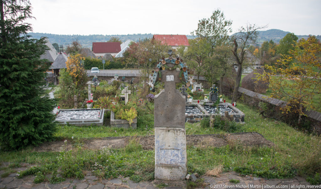 Hillside cemetery with grave plots and crosses overlooking village houses and wooded ridges in autumn.