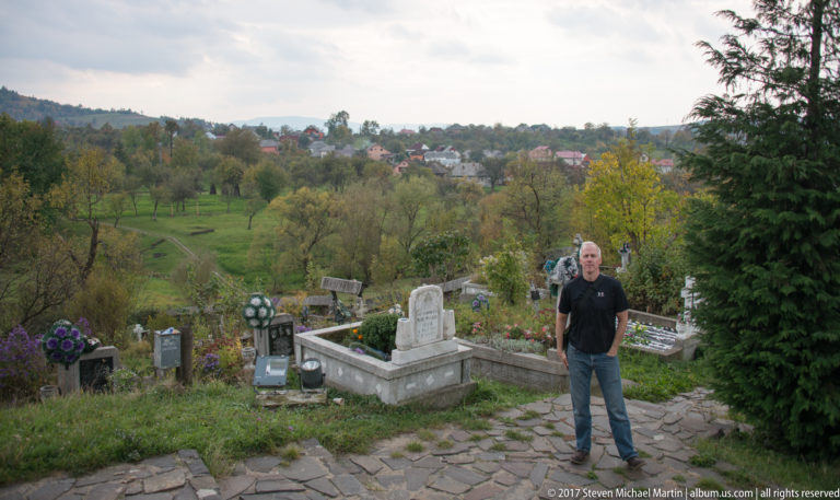 Man standing on a stone cemetery path with graves, orchards, village houses, and autumn hills behind him.