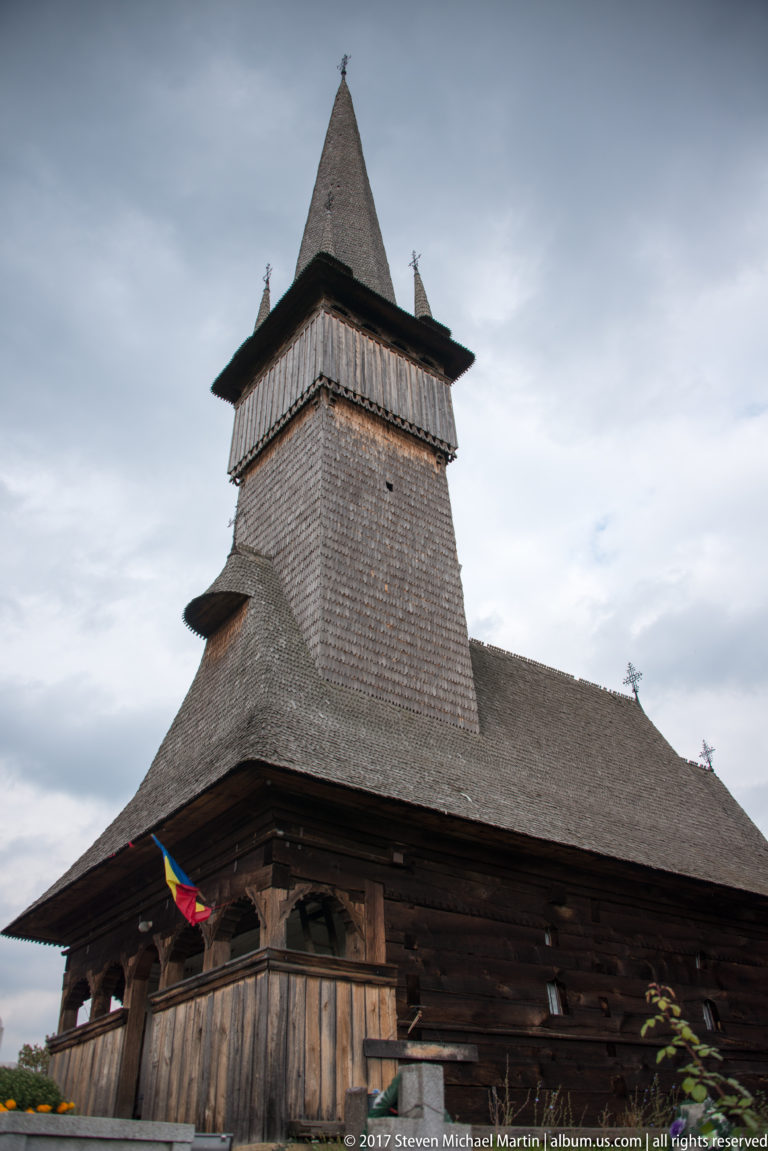 Low-angle view of a dark wooden church with broad shingled bell tower, deep eaves, and Romanian flag.