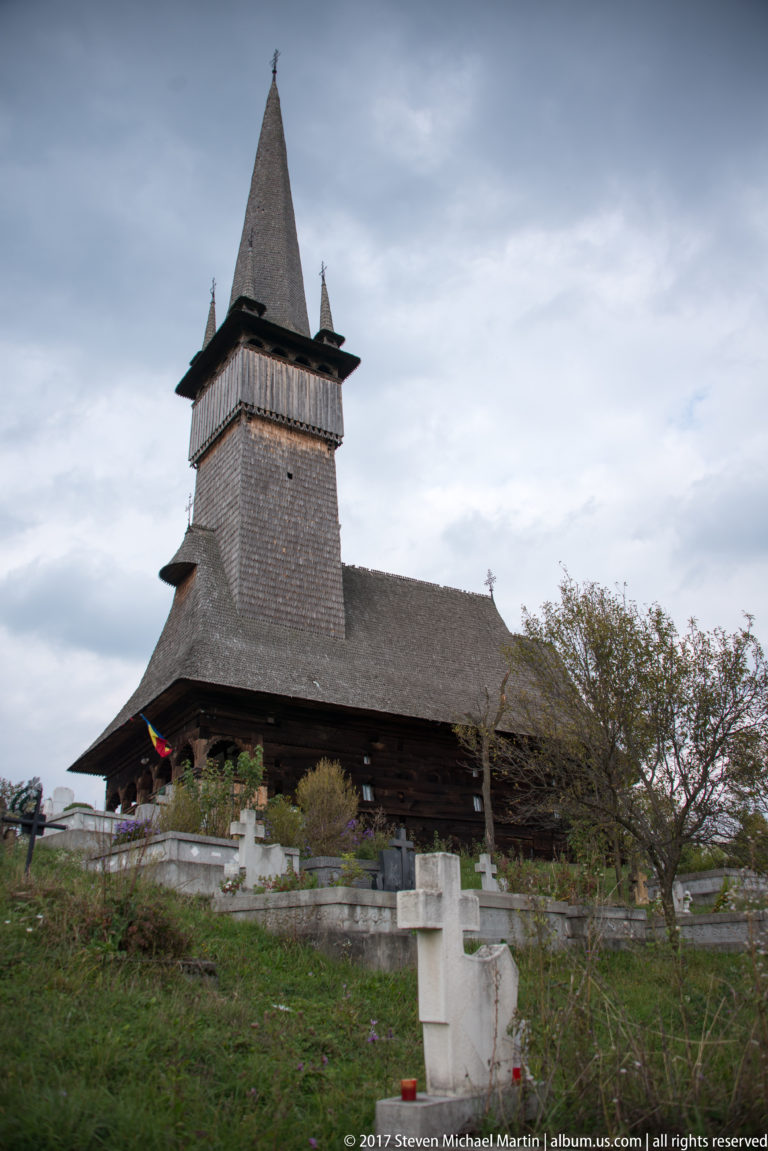 Dark wooden church with broad shingled tower above a sloping cemetery filled with grave crosses.