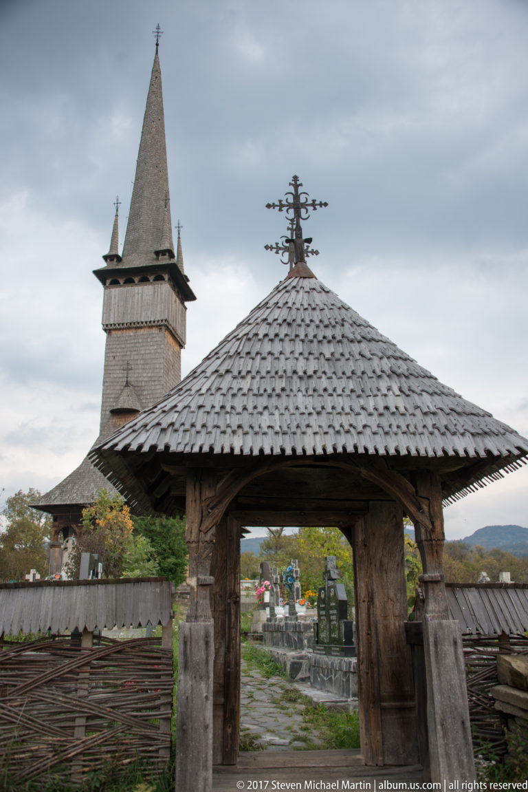 Roofed wooden churchyard gate with carved posts, shingled roof, woven fence, cemetery, and church tower beyond.