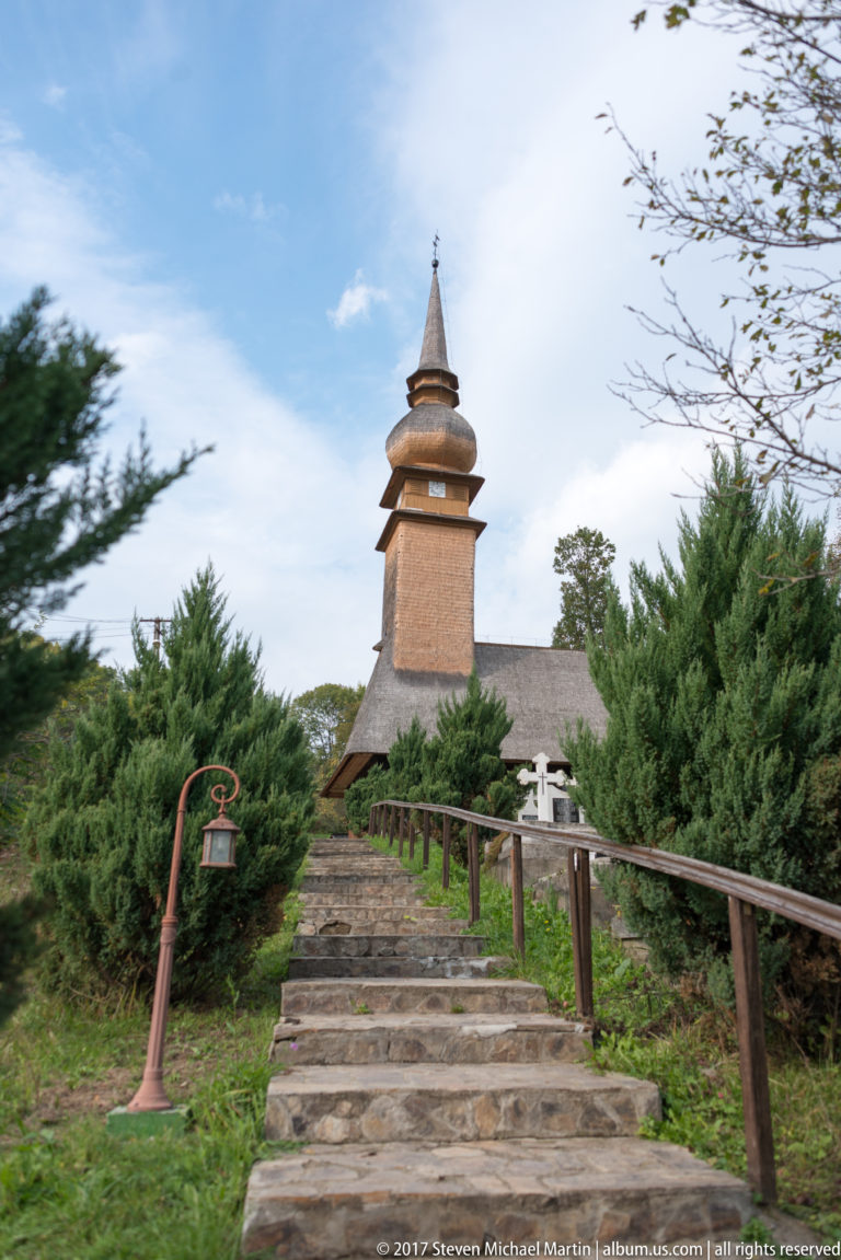 Stone steps and railings climbing through shrubs toward a wooden church with tiered shingled tower and pointed spire.