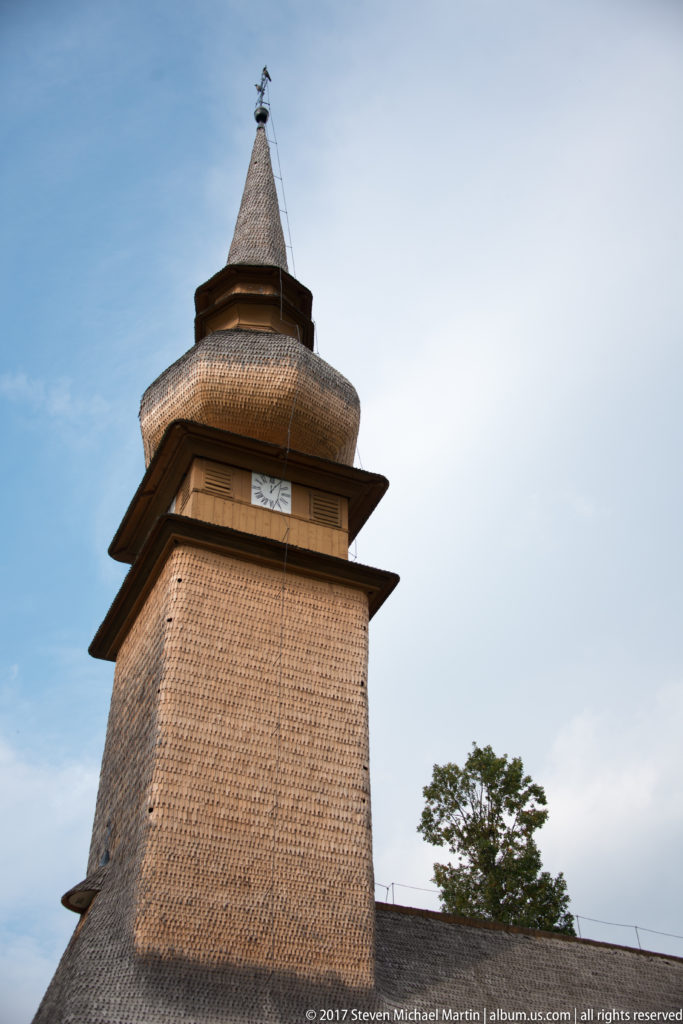 Close low-angle view of a square wooden church tower covered in shingles and capped by layered flared roofs.