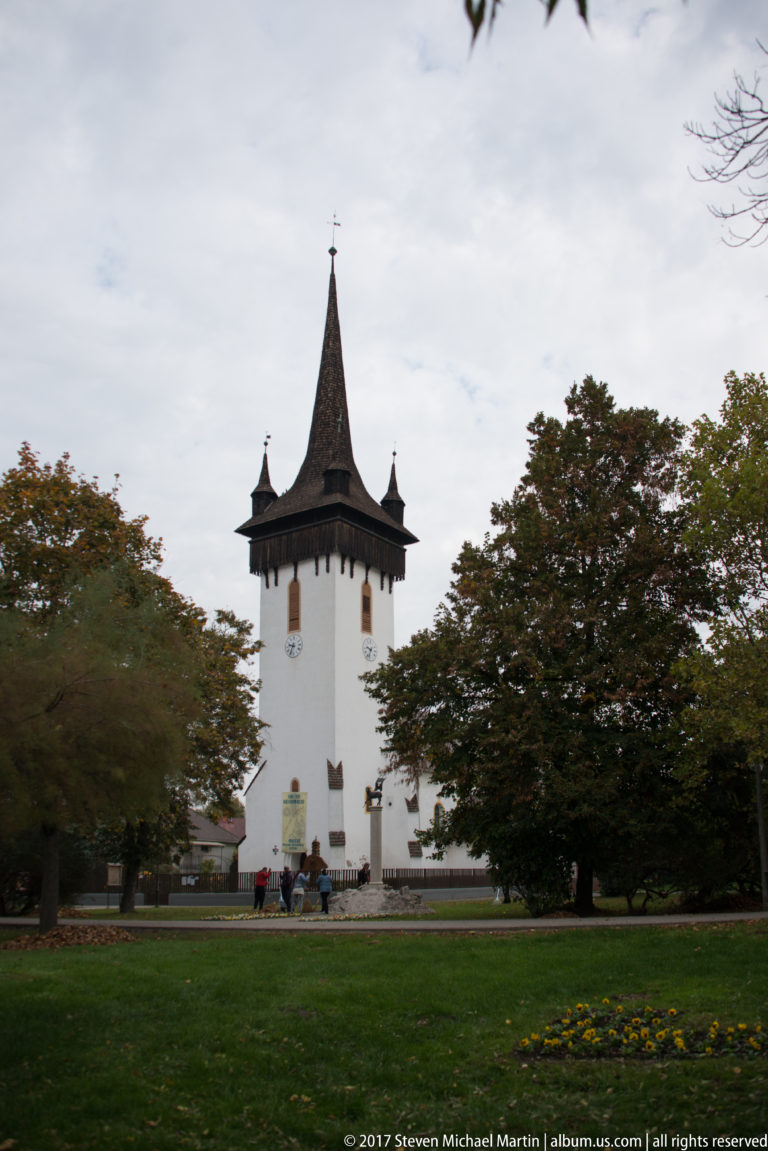 White church with a steep shingled tower and four corner turrets beyond a broad lawn and mature trees.