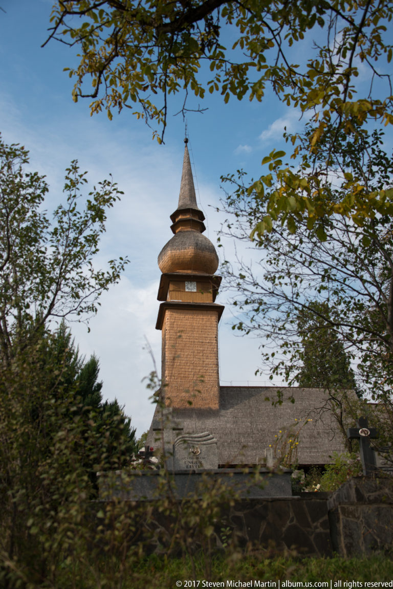 Wooden church behind trees and stone wall, with long shingled nave and tall tiered square tower.