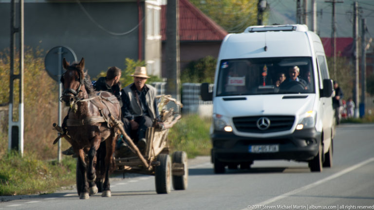 Two men riding a horse-drawn cart beside an approaching white minibus on a rural two-lane road.