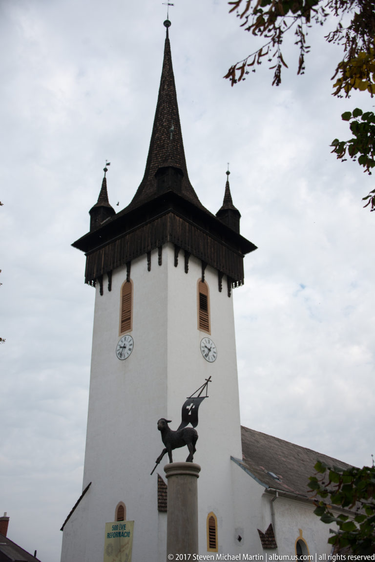 Close view of a white church tower with clocks, steep shingled spire, corner turrets, and a dark metal animal figure.