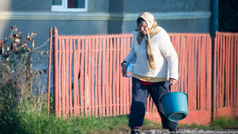 Elderly woman in a headscarf carrying a blue bucket beside a red wooden village fence.