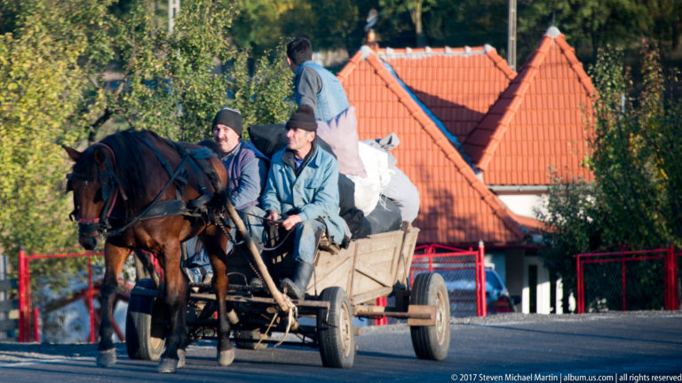 Three men riding a wooden horse-drawn cart loaded with white sacks along a village road.