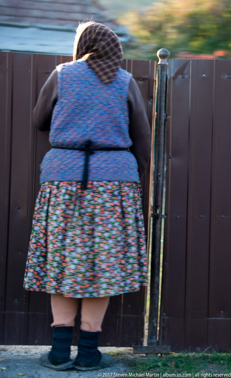 Woman seen from behind in patterned skirt, blue vest, head covering, and heavy socks beside a metal gate.