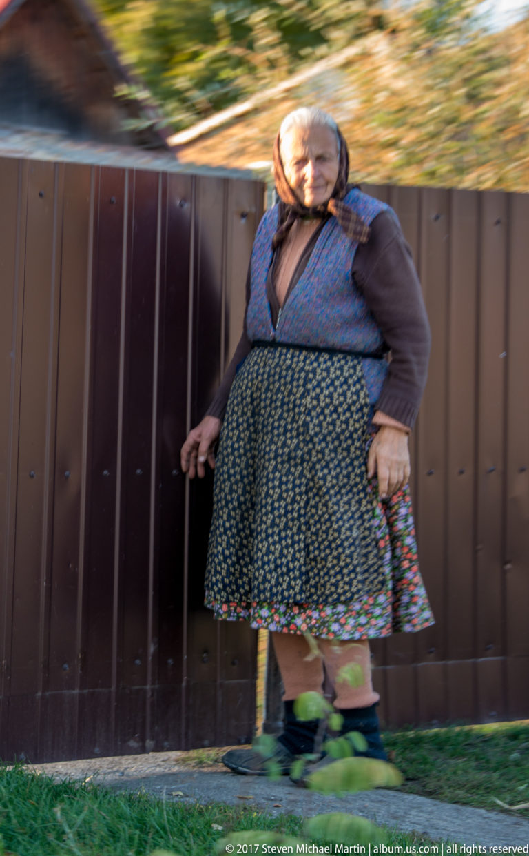 Woman in headscarf, blue vest, floral skirt, and green leg wraps beside a dark village fence.