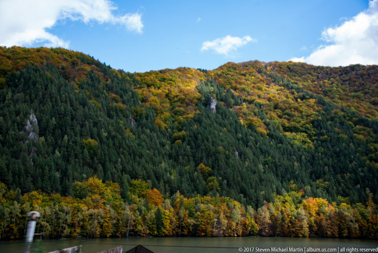 Autumn-covered mountain slope with dark conifer stands, small rock outcrops, and a narrow strip of water below.