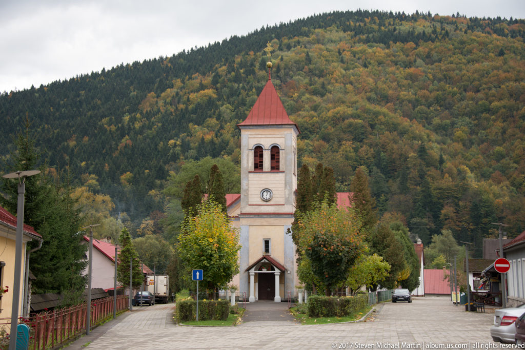 Small village church with square bell tower and red roof at the end of a street below wooded hills.