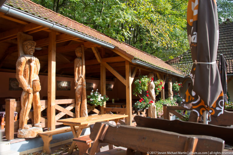 Carved wooden figures, benches, flowers, and timber posts beneath a covered outdoor terrace.