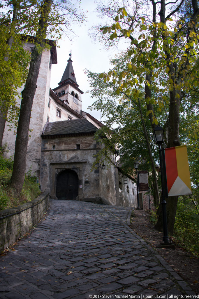 Shaded cobbled lane through trees leading uphill toward Orava Castle’s gate complex.