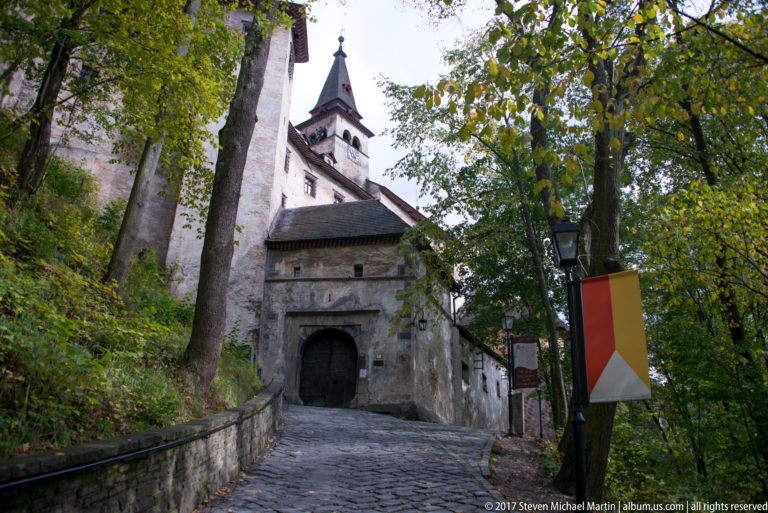 Cobbled uphill path between stone walls leading to a roofed gate and tower at Orava Castle.
