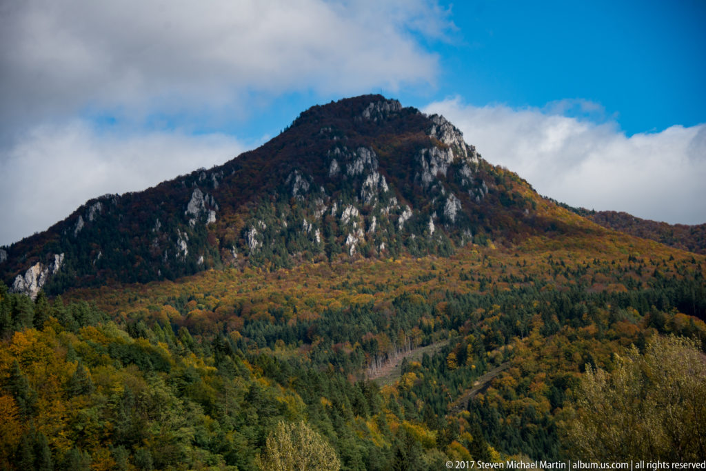 Rocky forested mountain ridge covered with autumn foliage beneath blue sky and clouds.