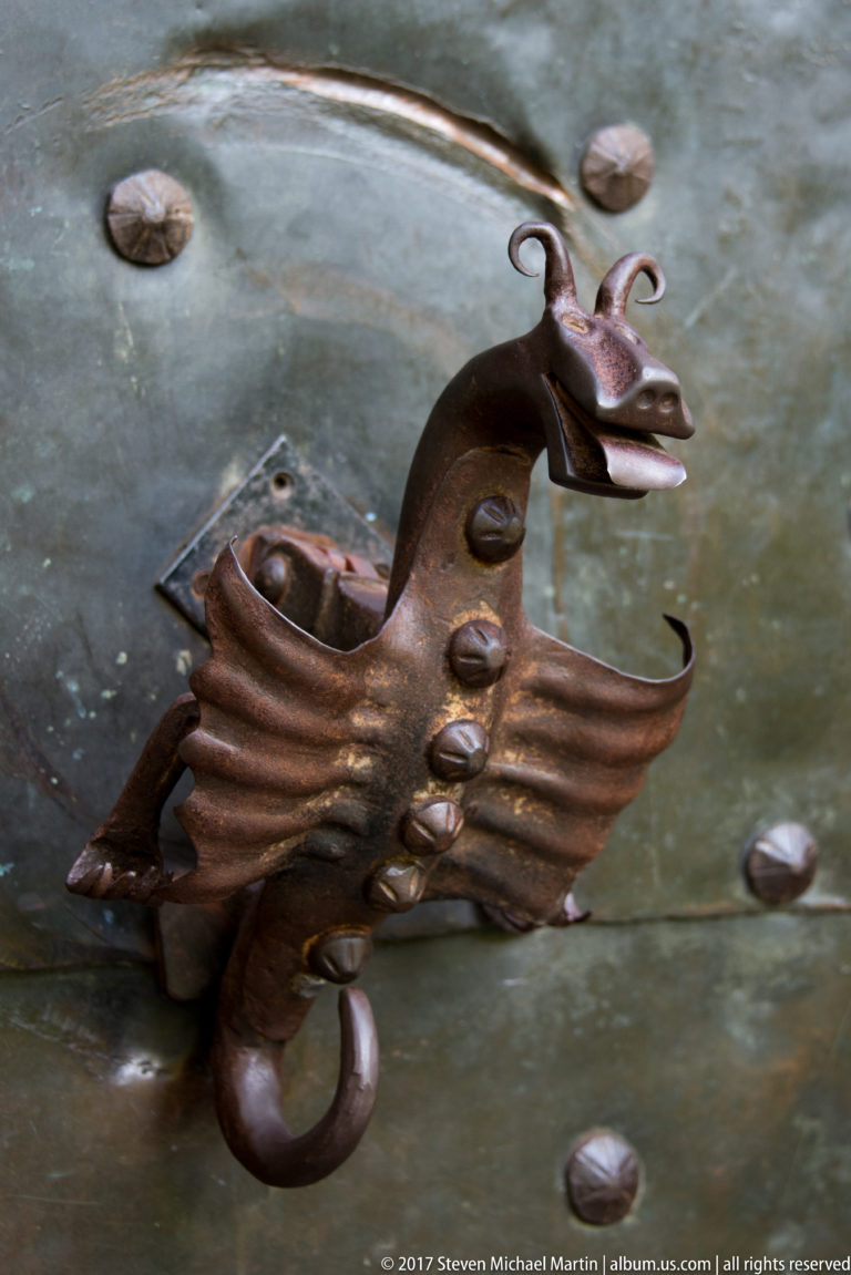 Winged dragon-shaped iron fitting with curled tail mounted on a riveted metal door.