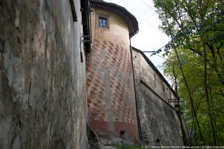 Rounded weathered tower joined to a high outer wall of Orava Castle beside trees.