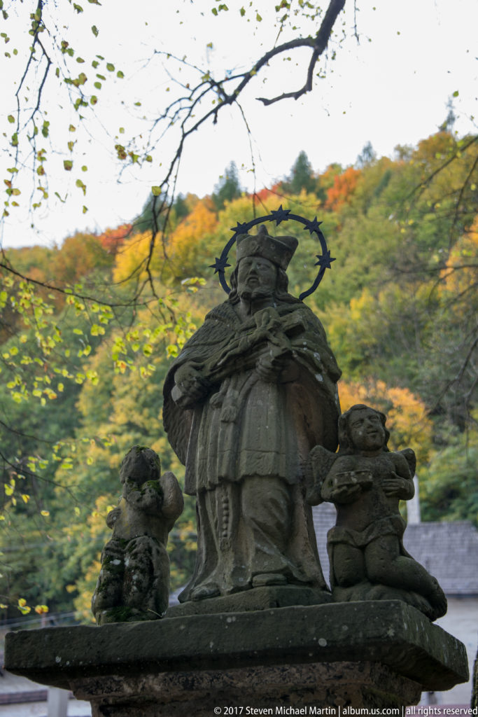 Weathered stone religious sculpture with haloed central figure and two smaller figures against autumn woodland.