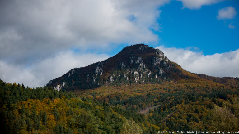 Forested mountain ridge with pale limestone outcrops and mixed autumn foliage beneath blue and cloudy sky.