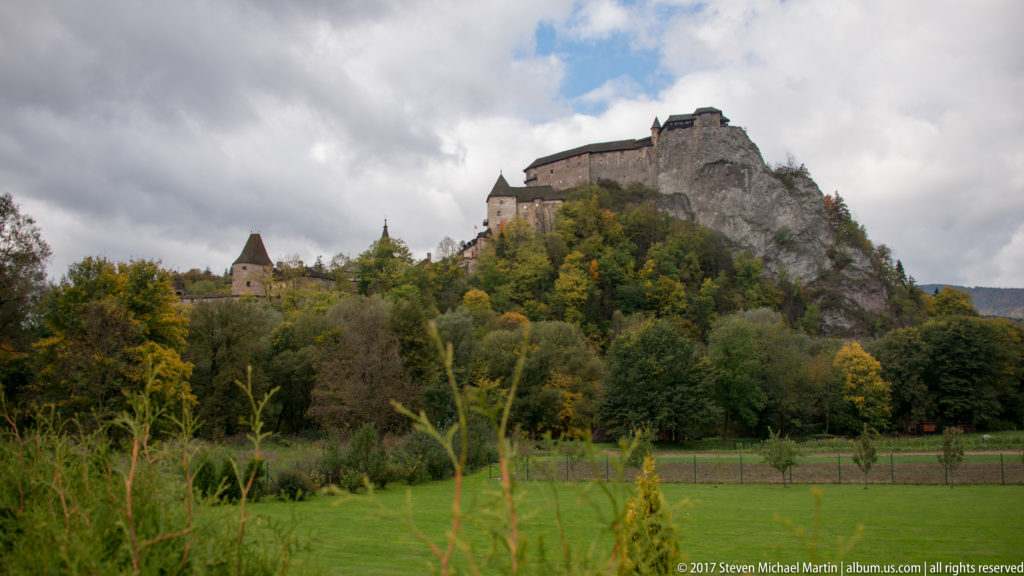 Orava Castle on a wooded rock beyond a green field under a partly cloudy sky.