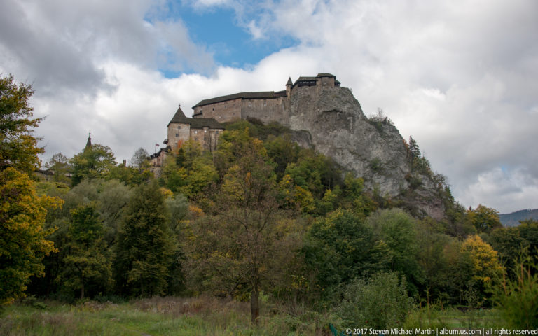 Distant view of Orava Castle on a rocky wooded ridge beyond meadow and autumn trees.