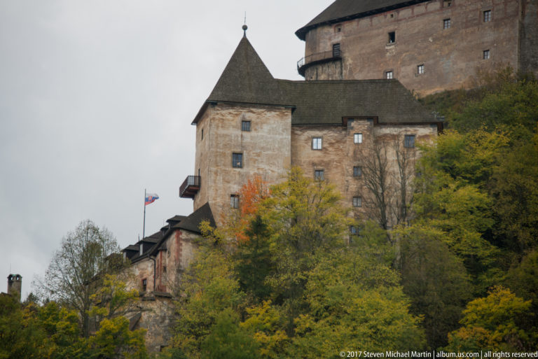 Clustered lower castle buildings and steep-roofed towers beneath a massive upper stone range.