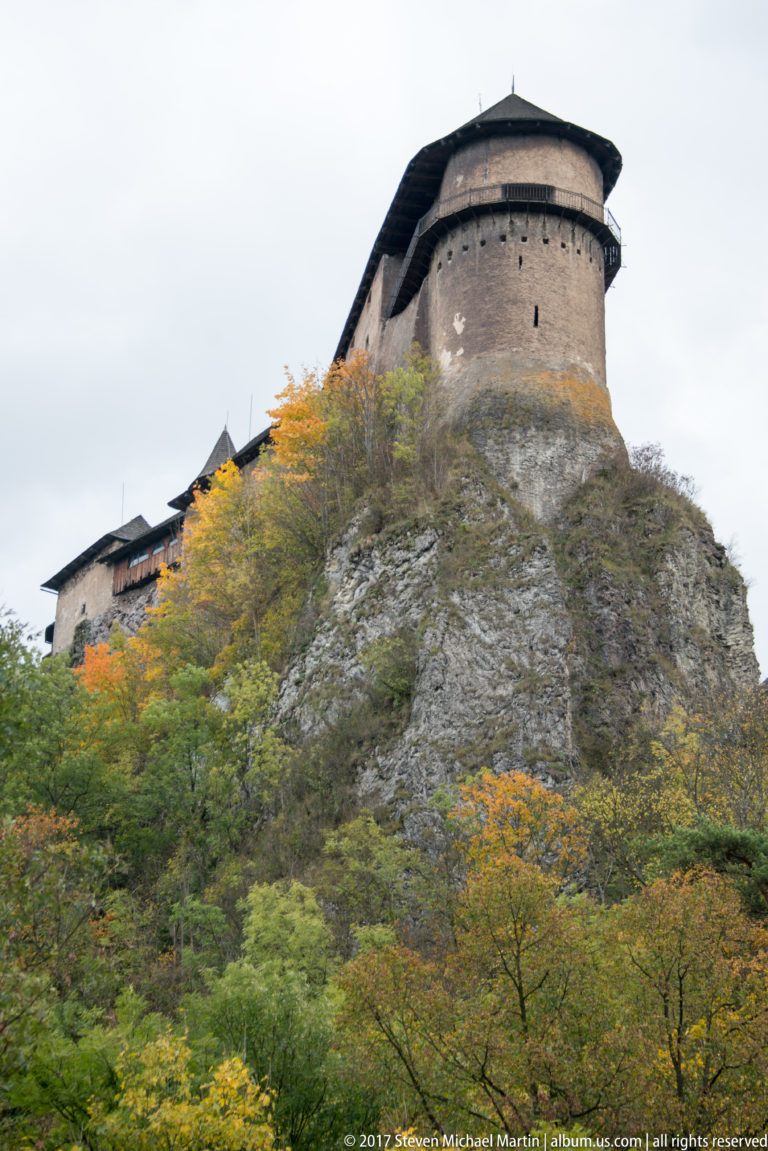 Rounded stone tower of Orava Castle above a bare rock face and autumn foliage.