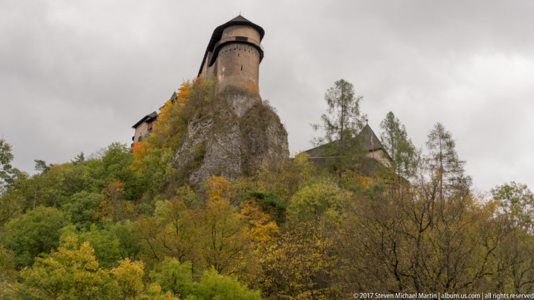Cylindrical castle tower with dark roof perched on a narrow rocky pinnacle above autumn trees.