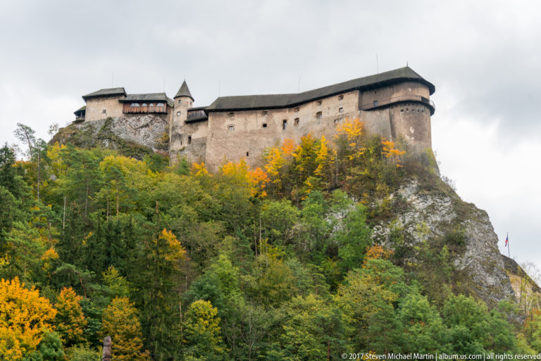 Long stone range of Orava Castle with round tower above bright autumn woodland and exposed rock.