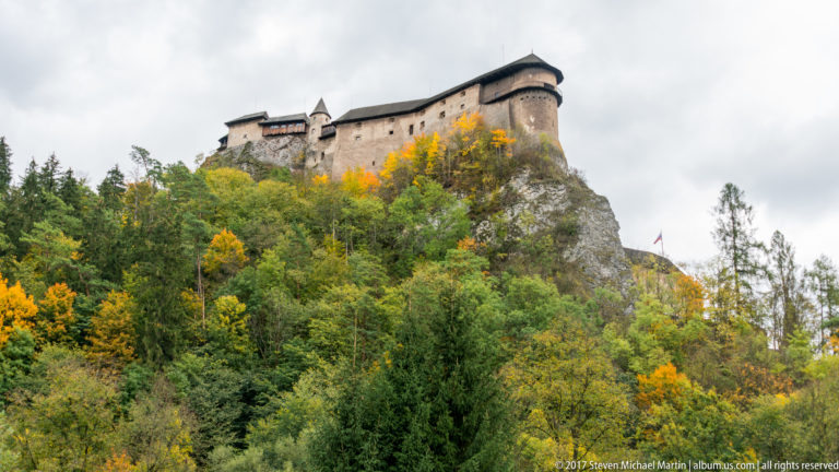 Upper walls and rounded tower of Orava Castle rising from a rocky outcrop above autumn trees.