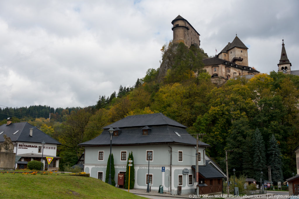 Orava Castle above roadside buildings in Oravský Podzámok, with wooded autumn slopes between town and fortress.