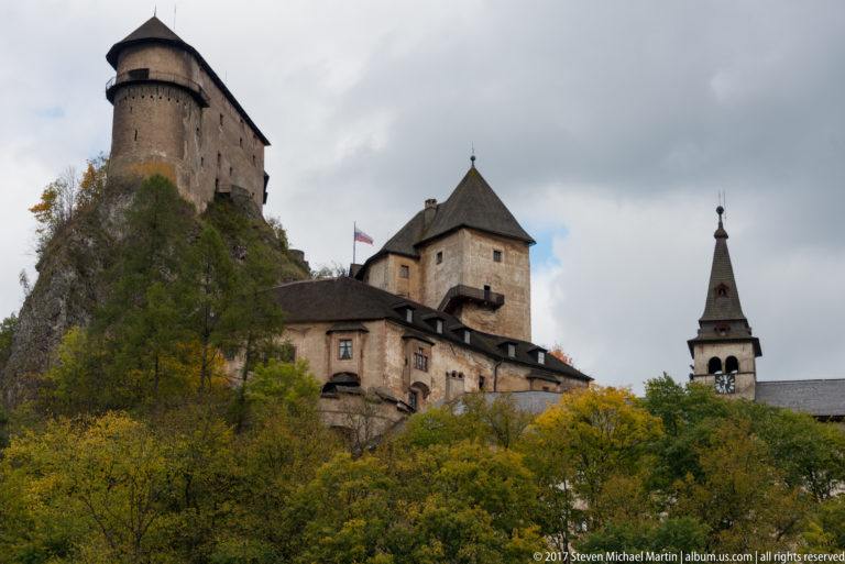 Cylindrical and square towers of Orava Castle rising above a wooded rocky slope in autumn.