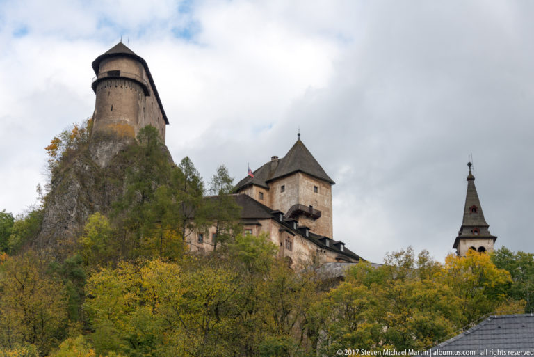 Orava Castle towers rising above dense autumn woodland, with a cylindrical tower at left and roofed towers to the right.