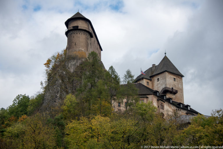 Cylindrical and square roofed towers of Orava Castle rising above autumn foliage on the rocky slope.