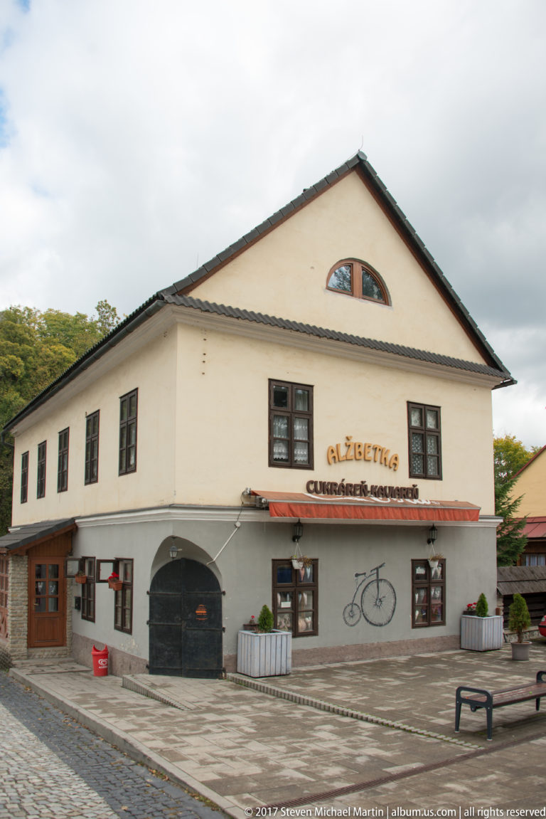Cukráreň Alžbetka façade with dark windows, central arch, planters, and bicycle motif.