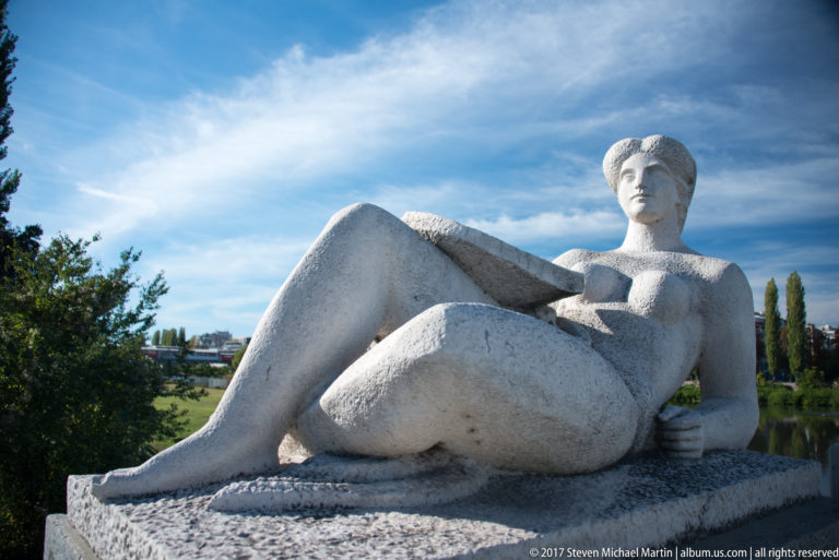 Large pale stone sculpture of a reclining female figure outdoors against trees, cityscape, and blue sky.