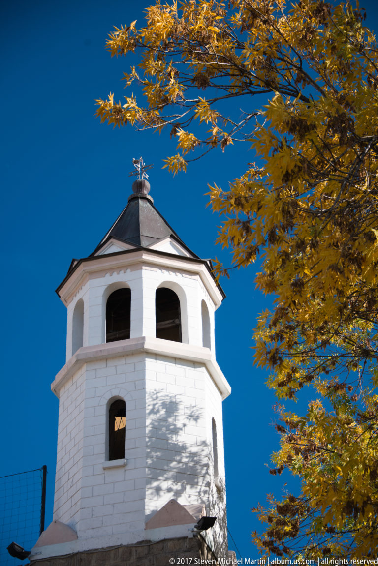White polygonal bell tower with arched openings and copper-colored roof, framed by golden leaves against blue sky.