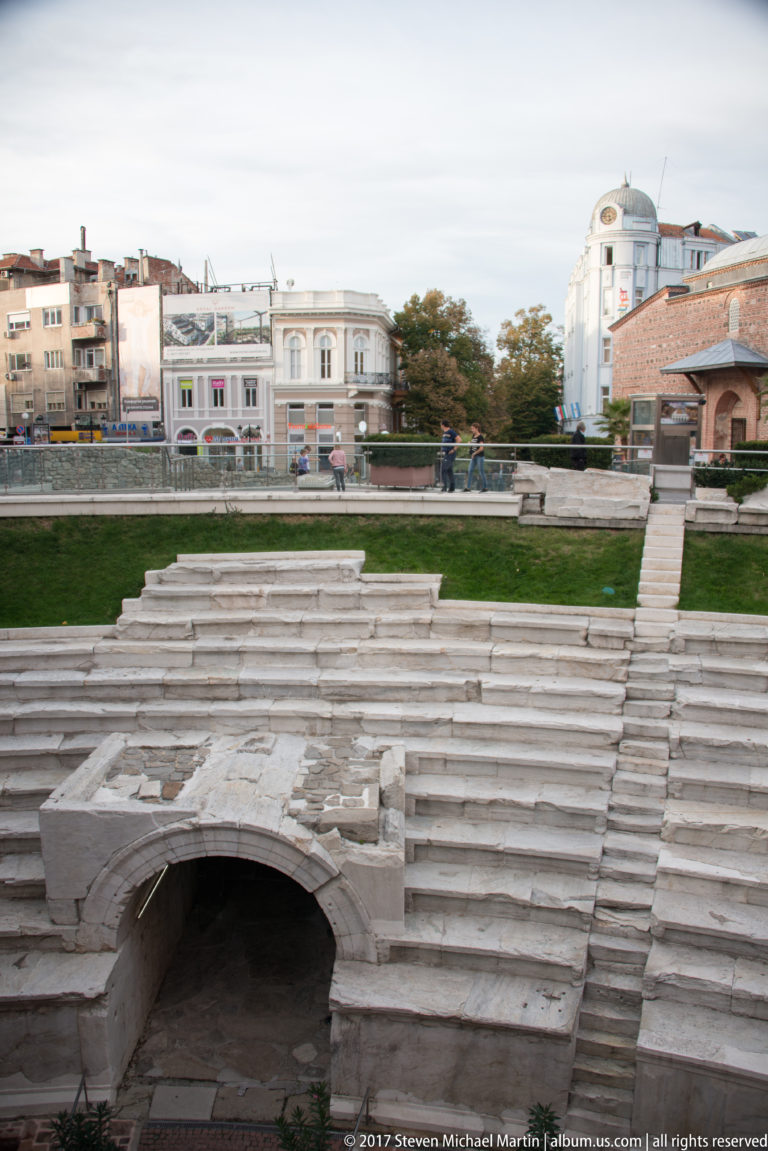 Curved stone seating and a vaulted passage at the Roman Stadium of Philippopolis, surrounded by modern central Plovdiv.