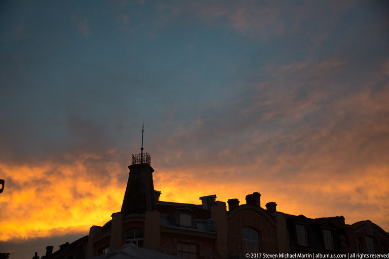 Dark silhouette of ornate Plovdiv rooftops and a pointed tower against an orange-and-blue sunset sky.