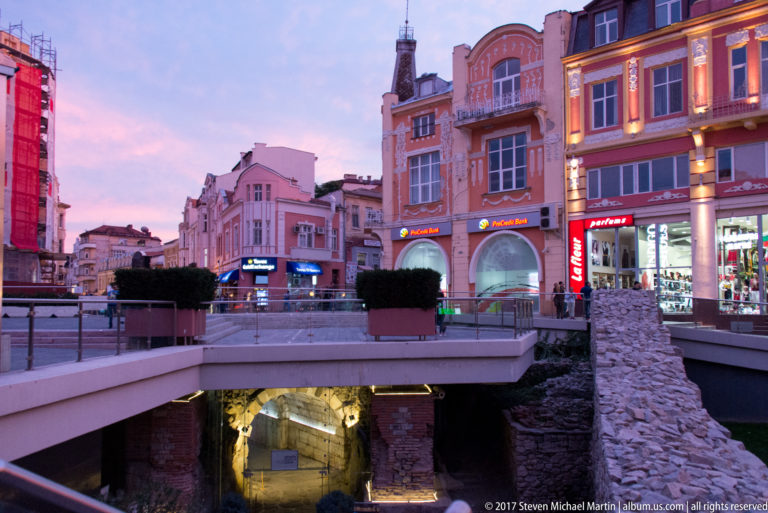 Illuminated Roman Stadium remains below a pedestrian street at dusk, framed by shops and historic façades in central Plovdiv.