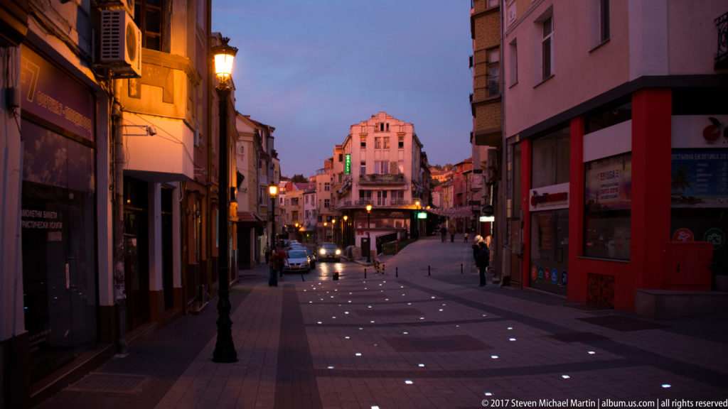 Pedestrian street at dusk with embedded pavement lights, warm shopfronts, scattered walkers, and buildings receding toward an intersection.