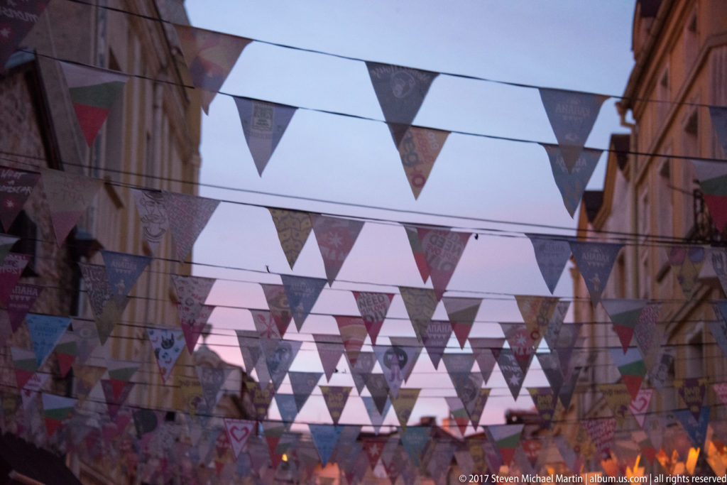 Multiple rows of printed triangular bunting suspended between buildings over a narrow Plovdiv street at dusk.