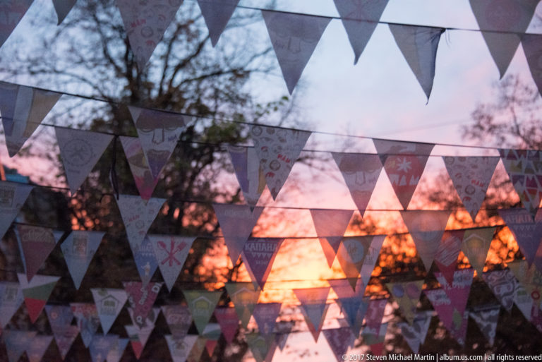 Overlapping triangular pennants backlit by an orange sunset, with tree branches and rooftops silhouetted behind.